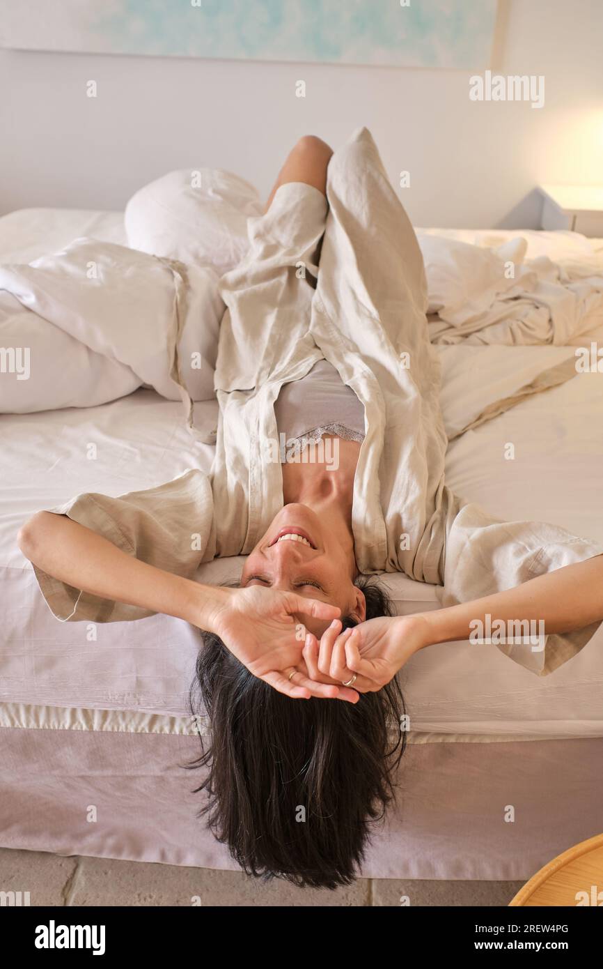 From above of cheerful female in sleepwear and bathrobe lying on bed upside down after awakening in morning with eyes closed Stock Photo