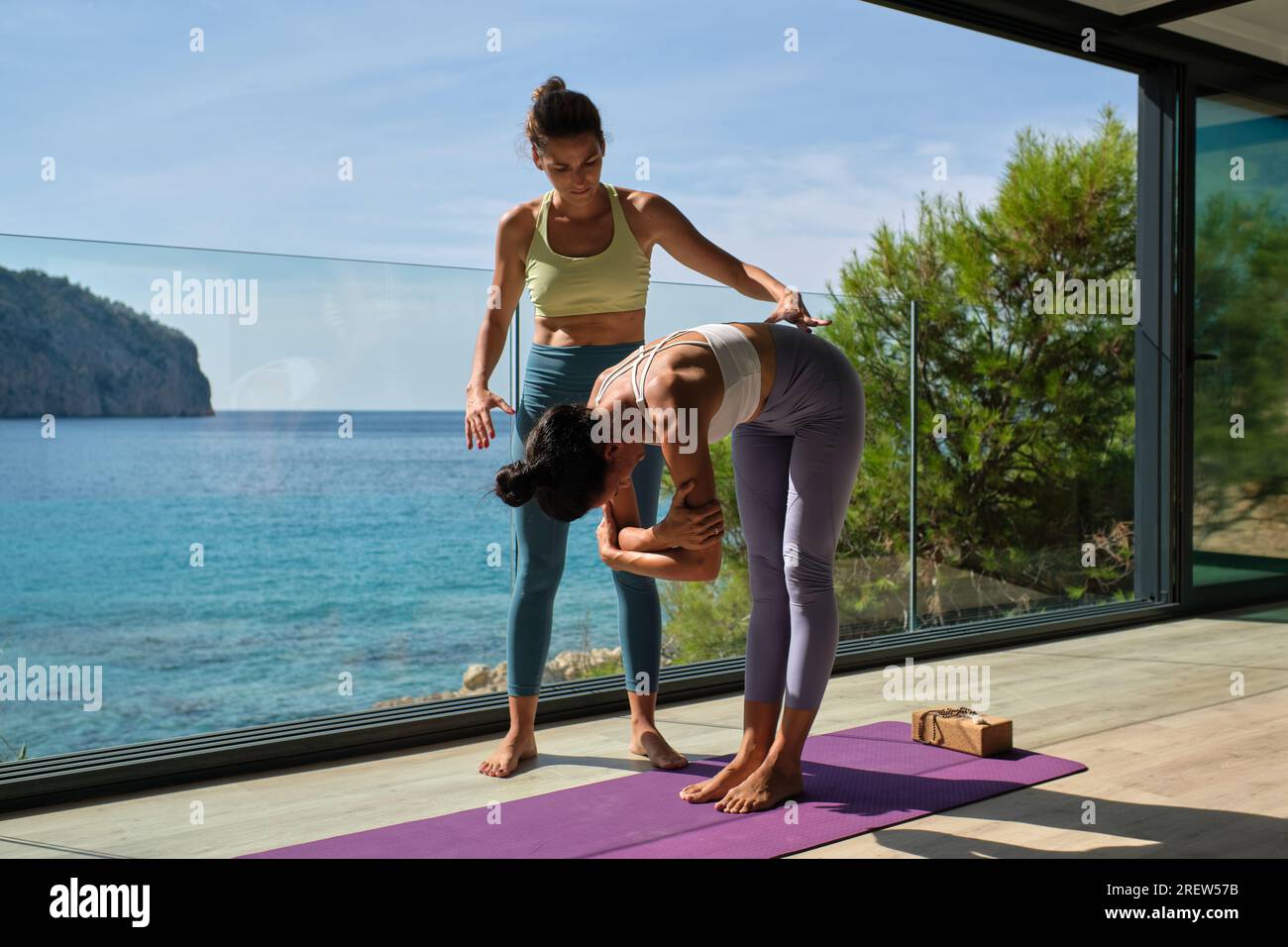 Full body of personal trainer assisting female bending forward with hands crossed while doing yoga on terrace of coast villa Stock Photo