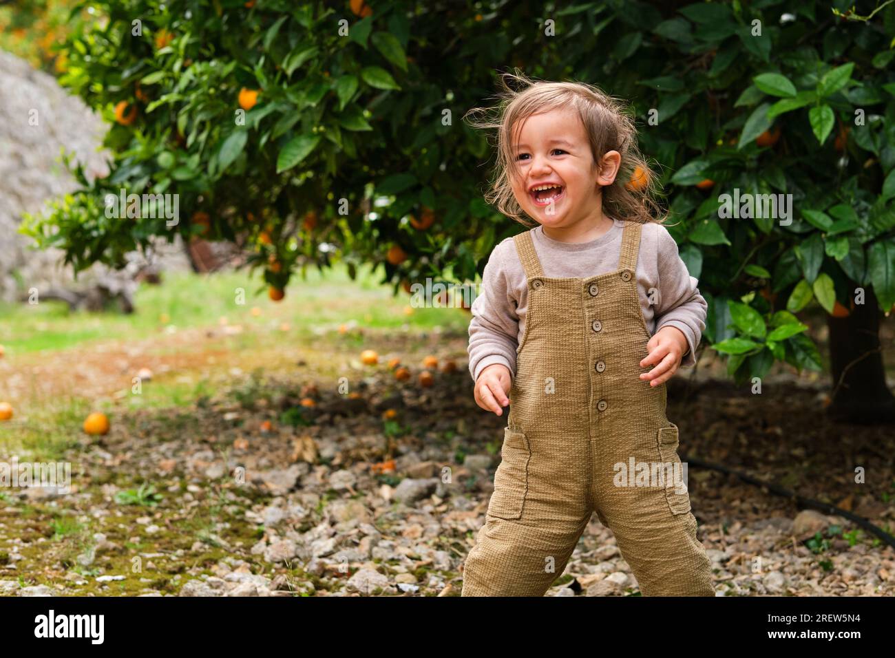 Content child in overalls laughing on pebble terrain and looking away against lush tree with oranges in countryside Stock Photo