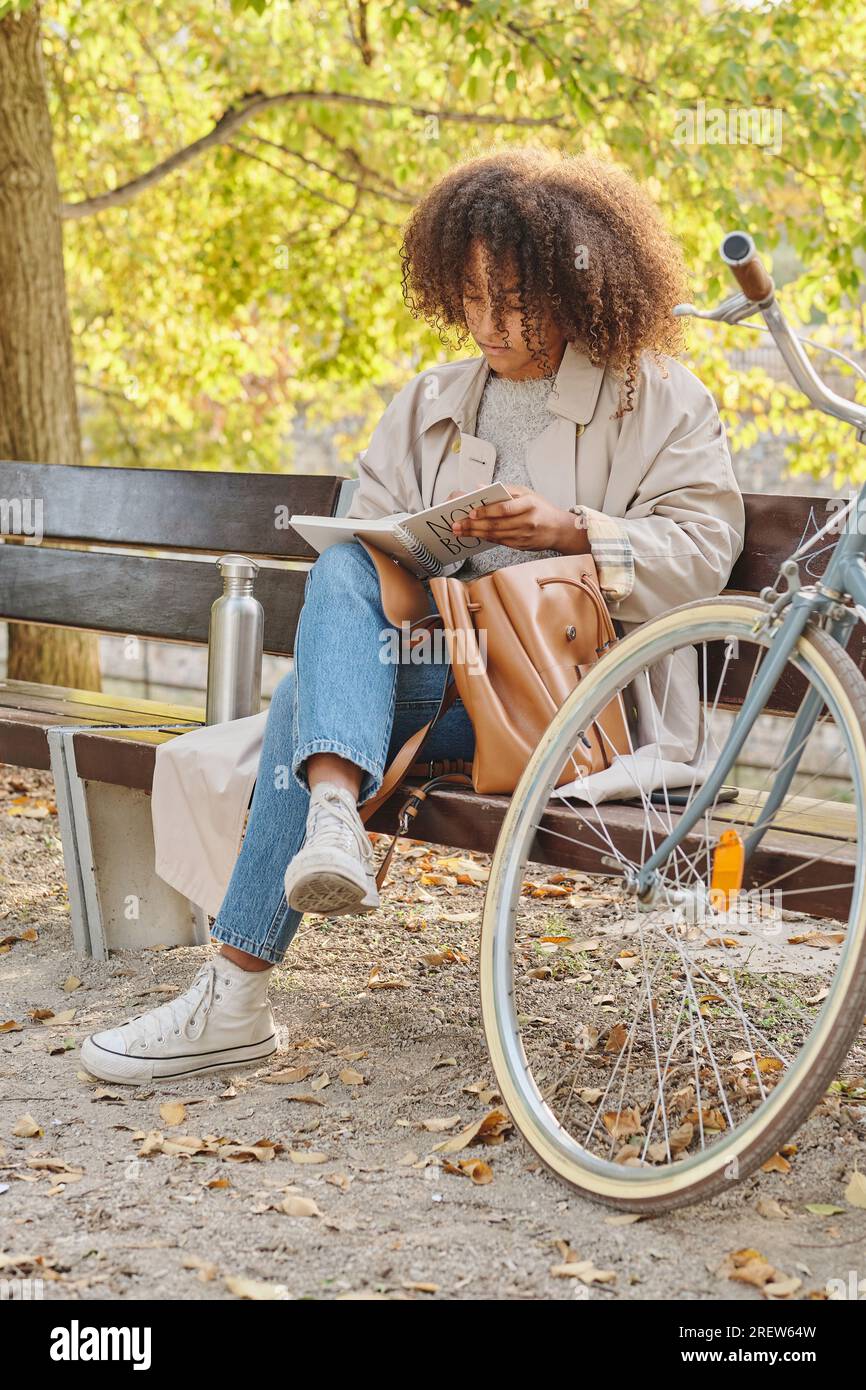 Full body of relaxed young African American female hipster in trendy outfit  sitting on wooden bench in green park in summer day Stock Photo - Alamy