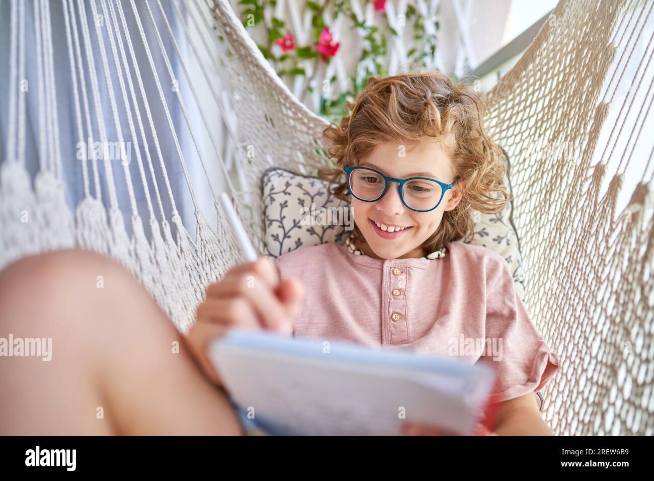 Smiling child in eyeglasses resting in knitted hammock with cushion and writing in diary with pencil on balcony at home Stock Photo