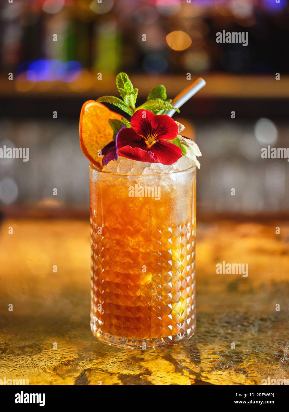 Long drink cocktail with ice cubes decorated with flower and dried orange slice served in modern bar on blurred background Stock Photo