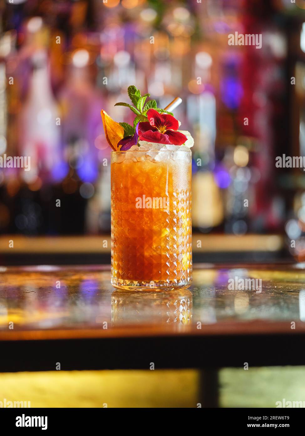 Alcoholic long drink cocktail decorated with flower and orange slice served with straw on bar counter against blurred background Stock Photo