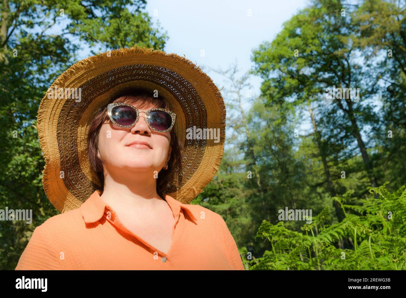 Middle-aged woman in orange dress, straw hat and sunglasses. In a summer shady green park. Place for text Stock Photo