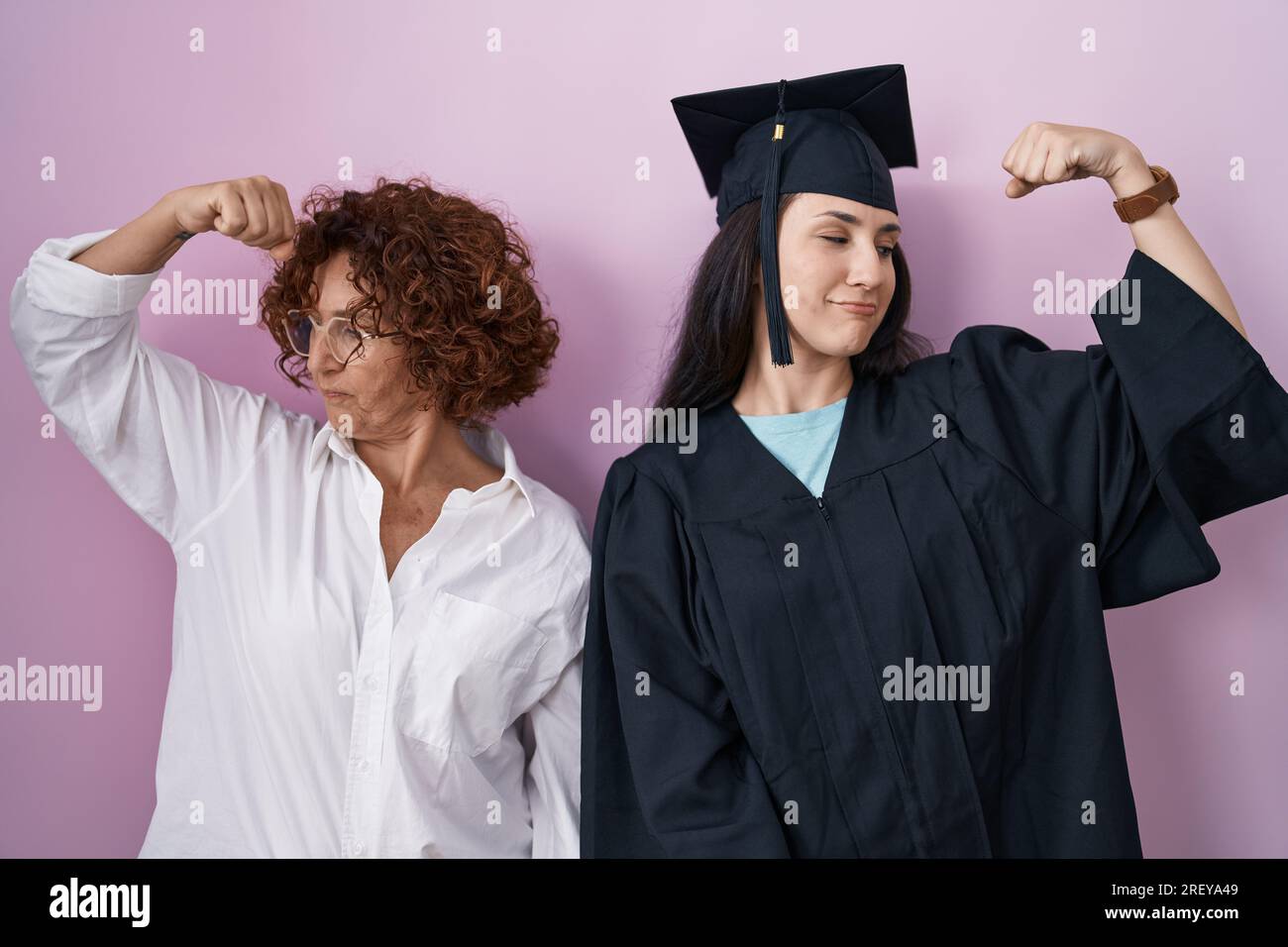 Hispanic mother and daughter wearing graduation cap and ceremony robe showing arms muscles smiling proud. fitness concept. Stock Photo