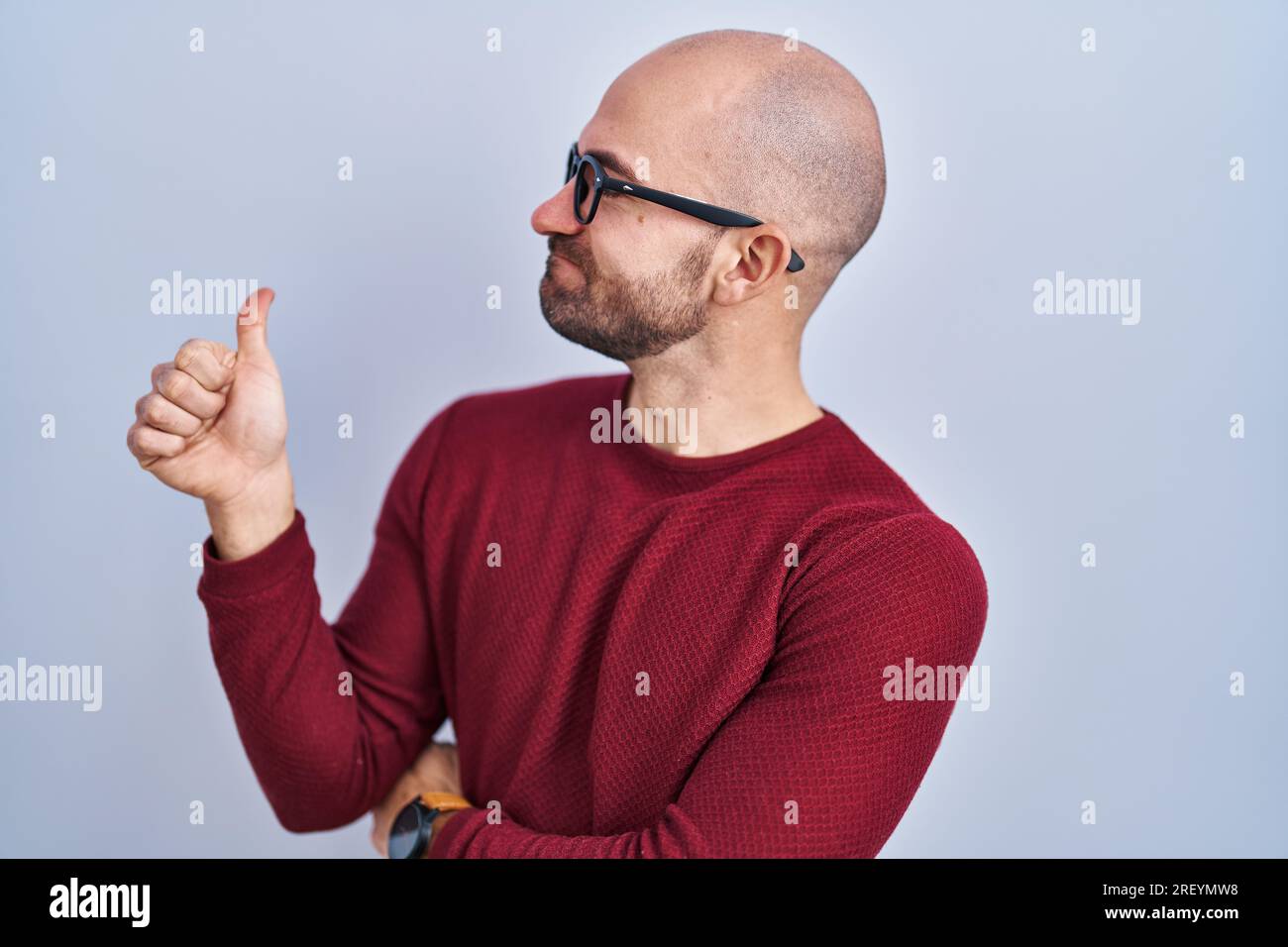 Young bald man with beard standing over white background wearing glasses looking proud, smiling doing thumbs up gesture to the side Stock Photo