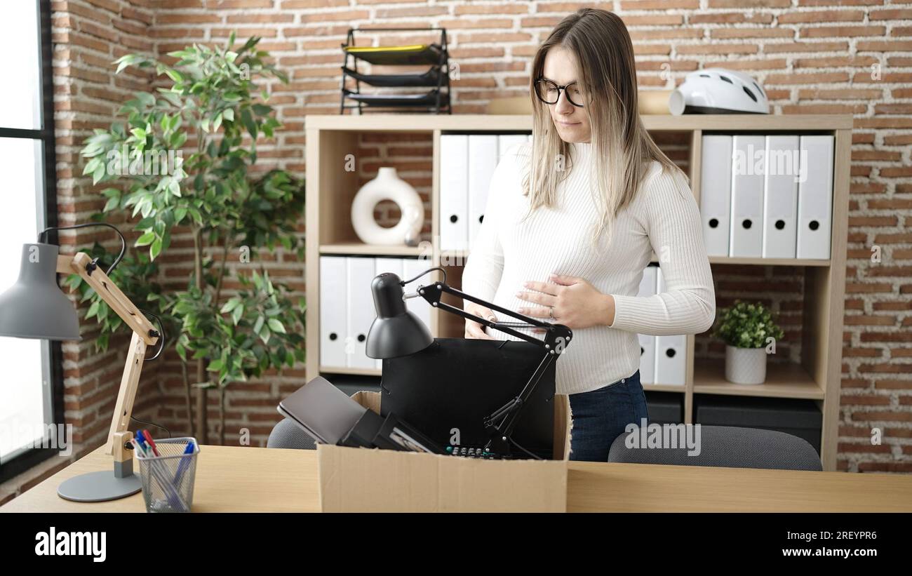 Young pregnant woman being fired packing belongings from workplace touching belly at office Stock Photo
