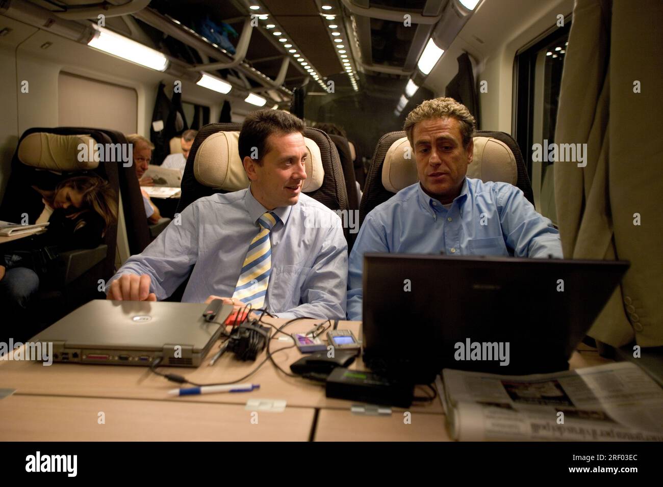 Two men in conversation on a train, using laptops and surrounded by passengers reading or resting. Stock Photo