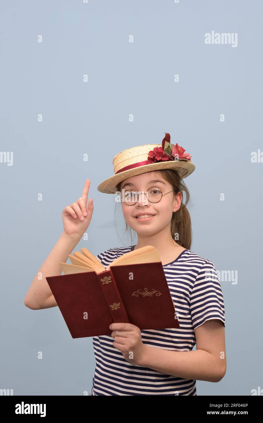 Teenage girl reading a book with enthusiasm while dressed in vintage clothing. a straw hat with flowers, glasses and a book in her hand. Stock Photo