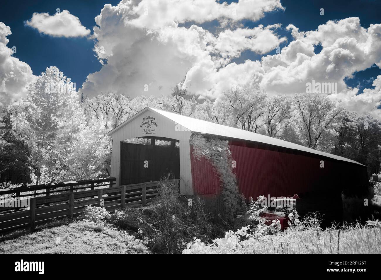 A colorized black and white image of a red covered bridge near Shieldstown, IN. White foliage with blue sky, red siding, and red water. Stock Photo