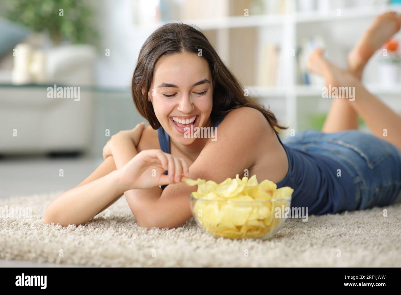 Happy woman laughing and eating potato chips on a carpet at home Stock Photo