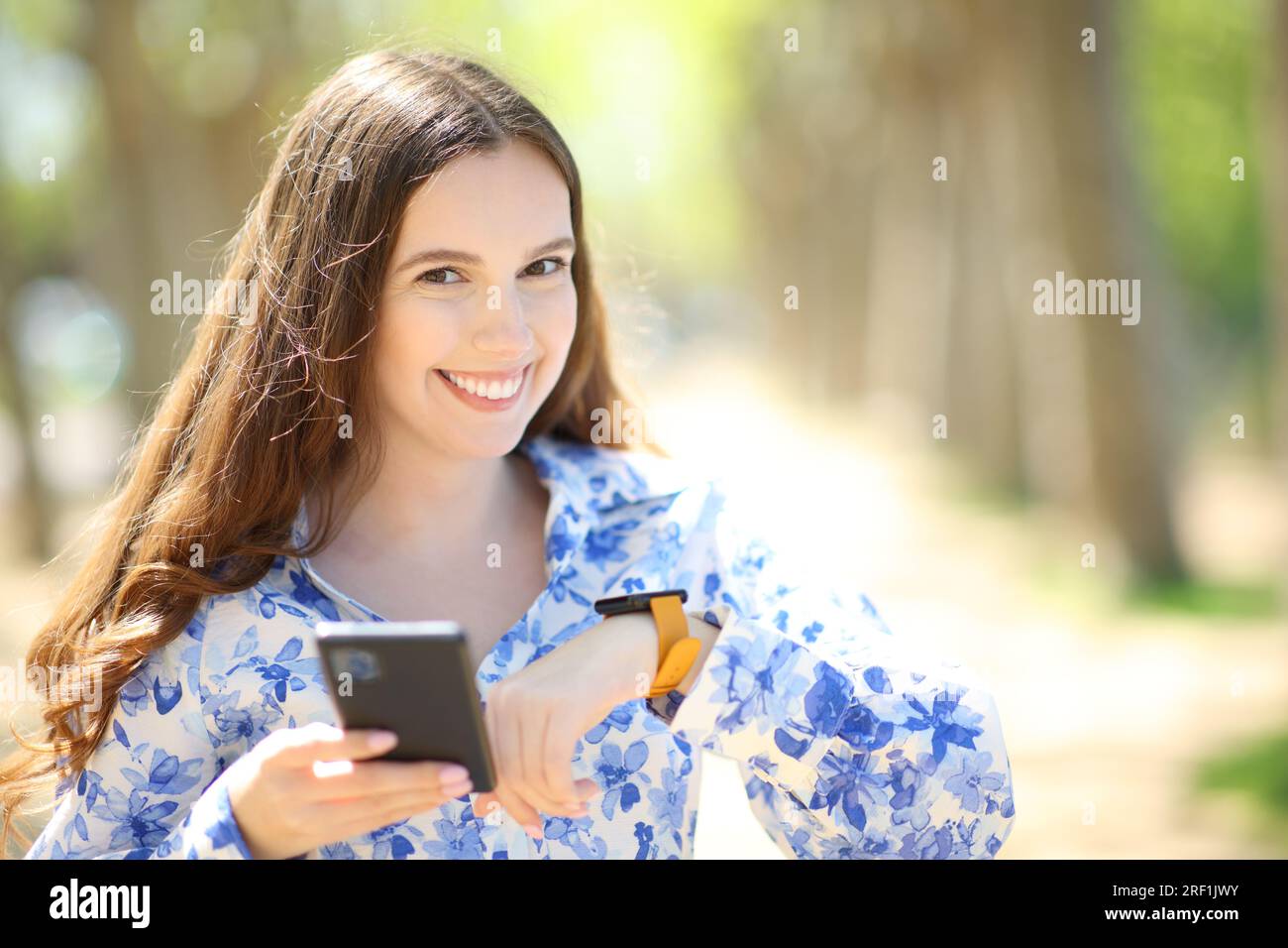 Happy woman wearing smartwatch holds phone looking at you  in a sunny park Stock Photo