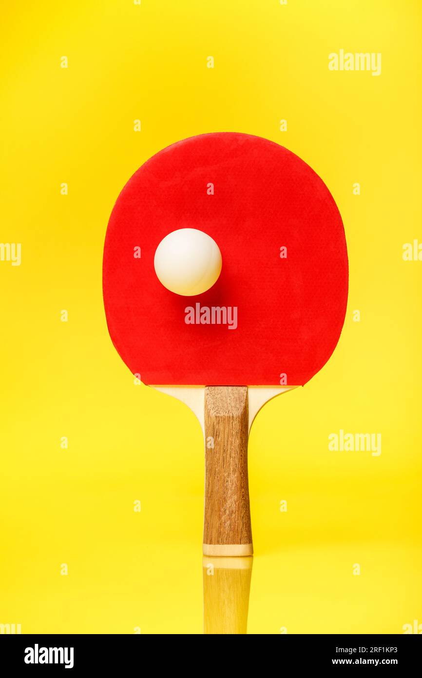 Red side of a ping pong racket with a white ball standing on a yellow background. TAble tennis