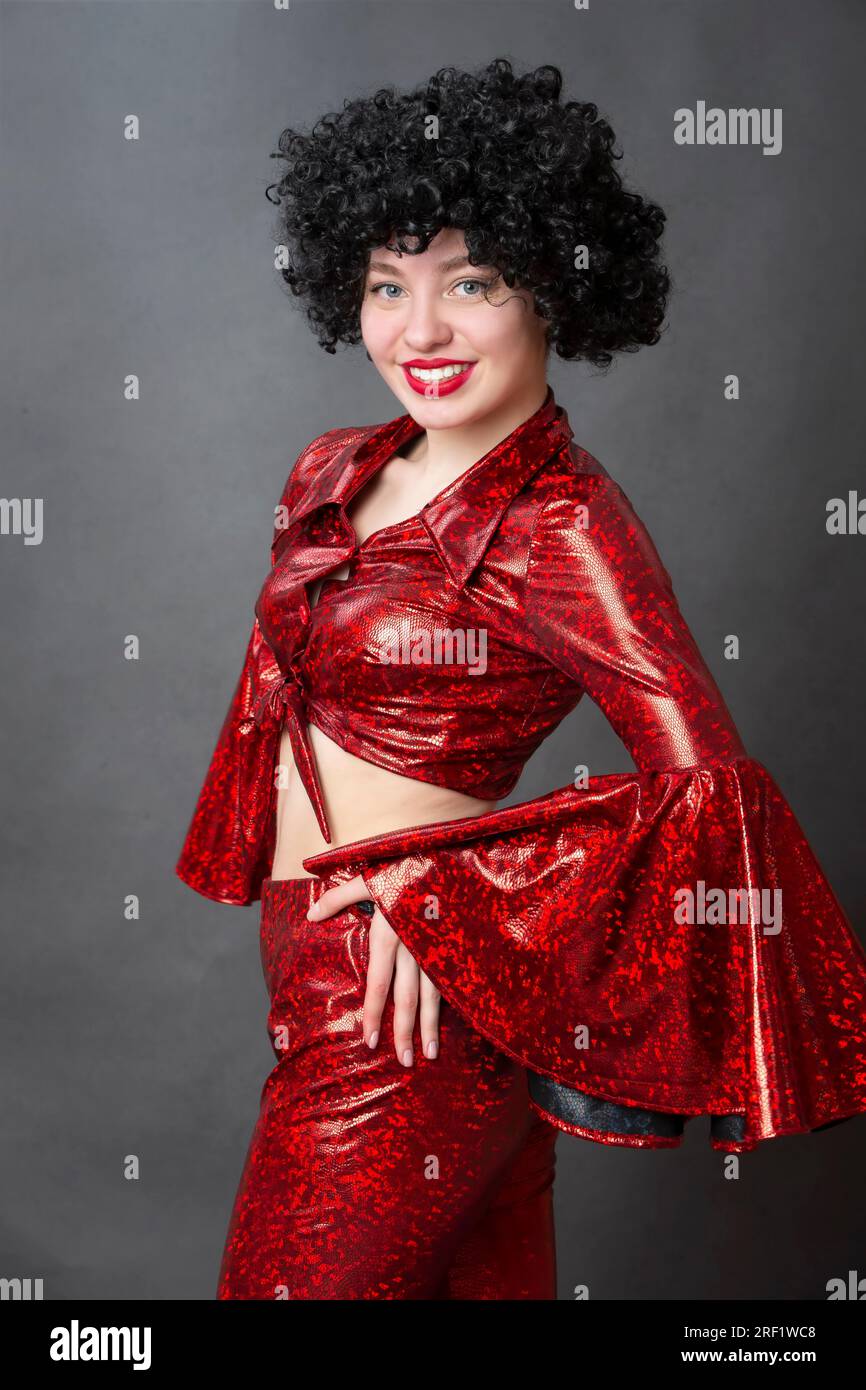 Vintage woman in afro wig and red disco costume on a gray background. Girl in the style of the seventies. Stock Photo