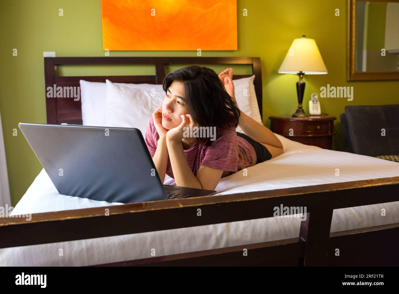 Young woman or teen lying down on bed using laptop computer, looking off to side bored or thinking Stock Photo