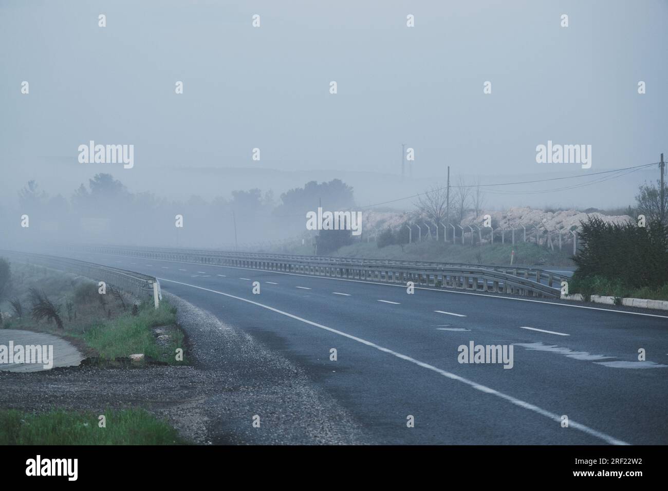 Dramatic scene resembling a movie frame, captured in the early hours of the morning. A winding road covered in thick mist, creating a low visibility a Stock Photo