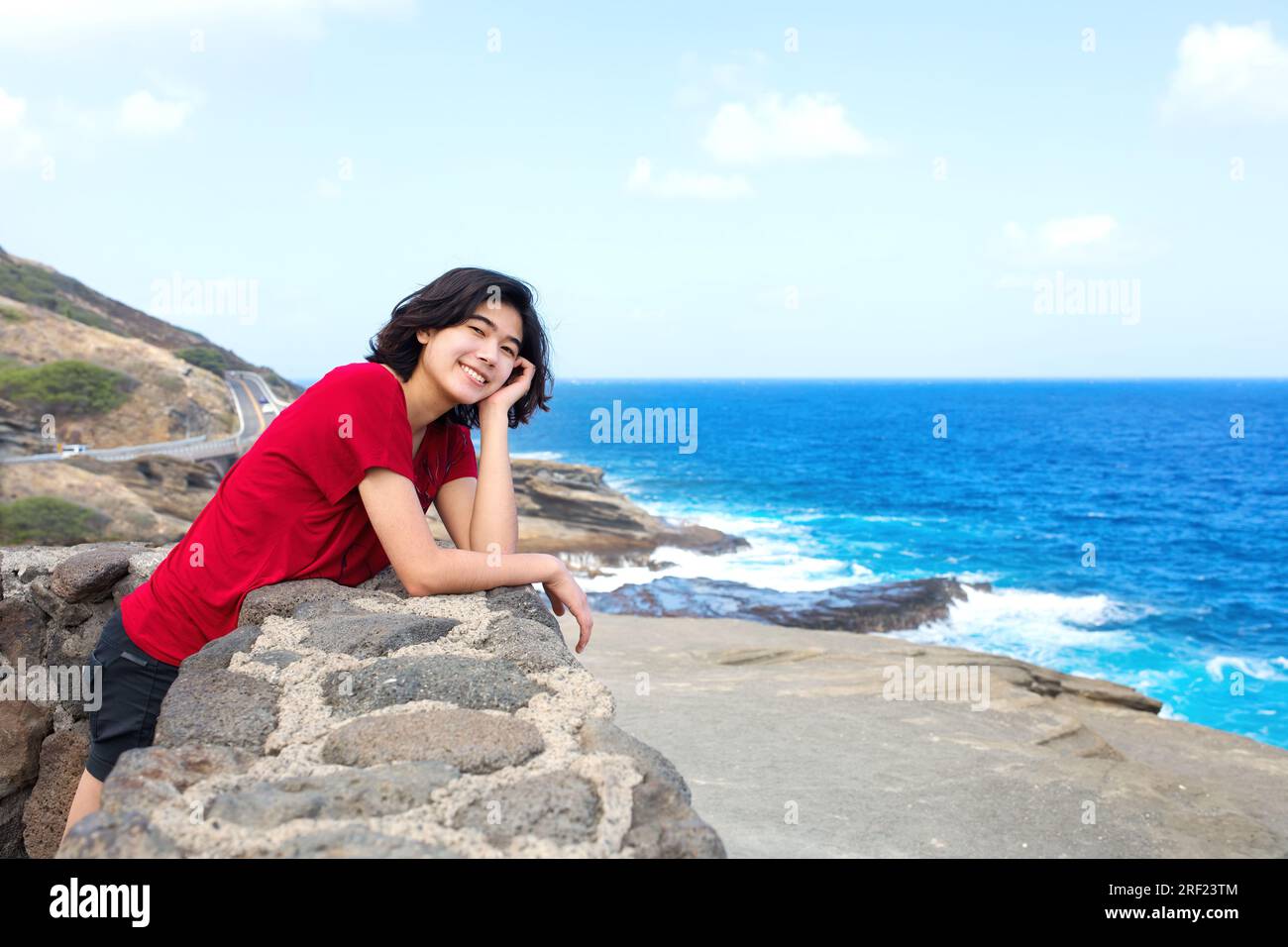 Young woman leaning against stone wall overlooking Hawaiian ocean from a high scenic viewpoint Stock Photo