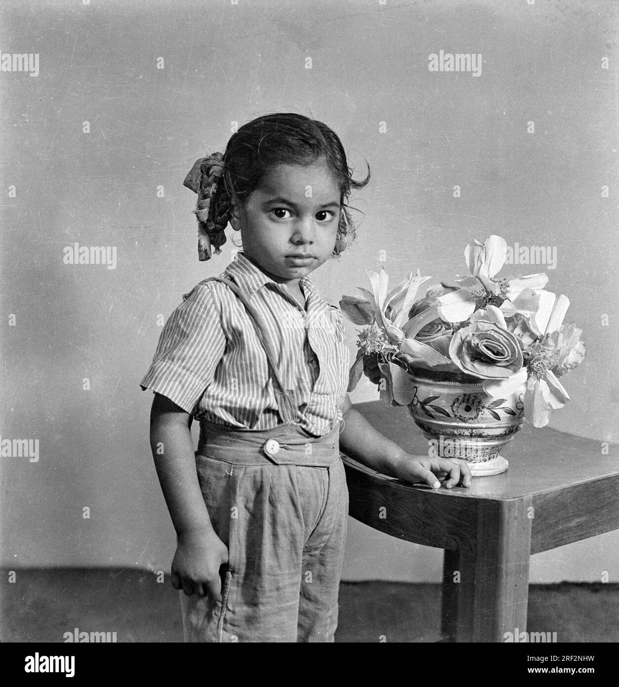 old vintage 1900s black and white studio portrait of Indian baby girl child wearing pant shirt standing artificial flowers stool India 1940s Stock Photo