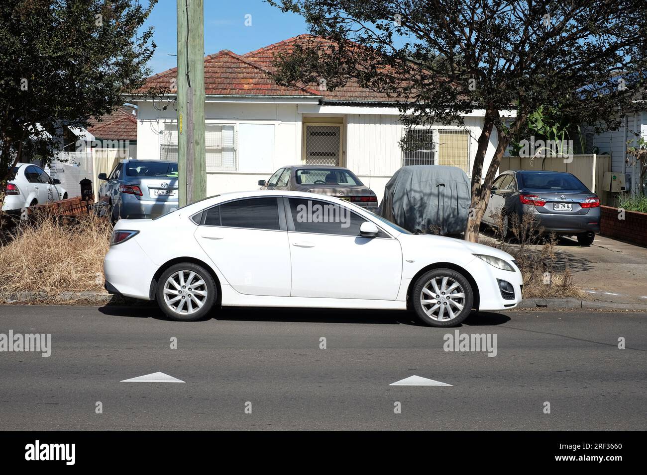 A single-story white Fibro house in suburban Caley Vale with 4 cars parked in the front yard & a white car on the street out front in Western Sydney Stock Photo