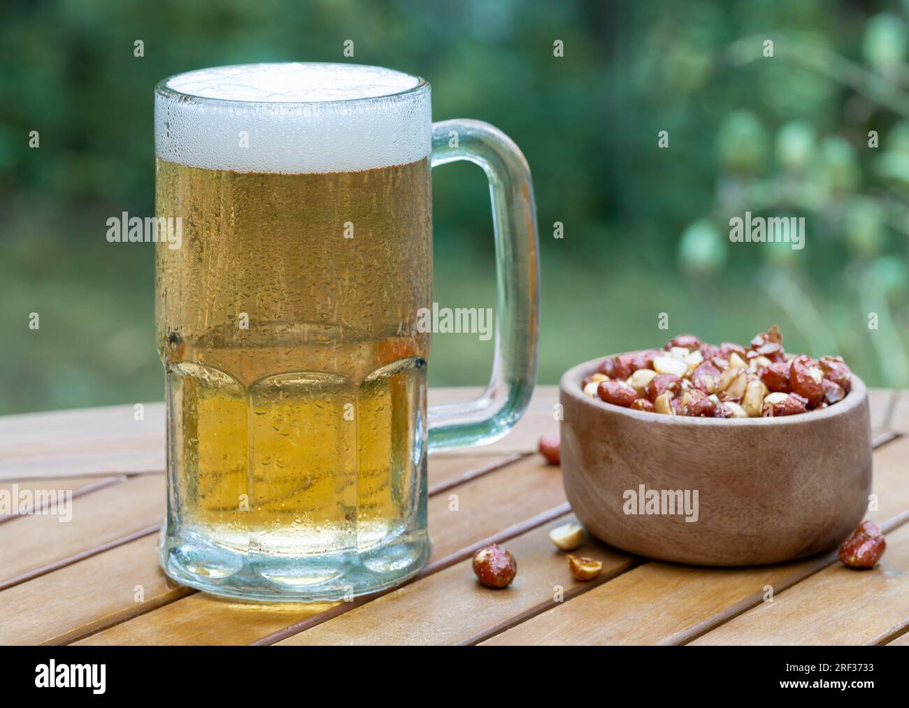 Glass mug of beer and bowl of nuts outdoors on wooden patio table with nature background Stock Photo