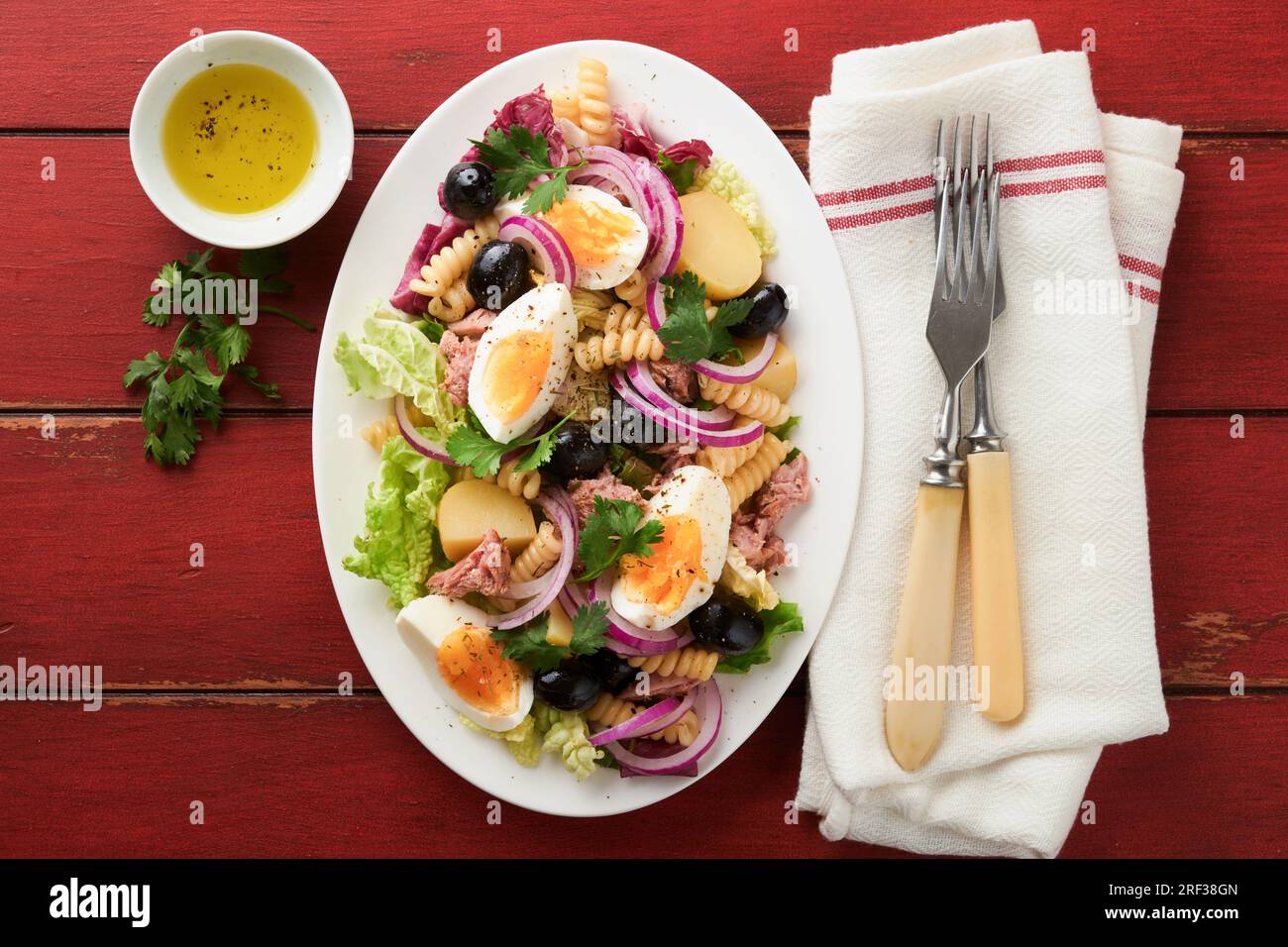 Tuna salad with pasta, eggs, potatoes, olives, red onions and sauce in white plate on old red rustic table background. Nicoise salad. French cuisine. Stock Photo