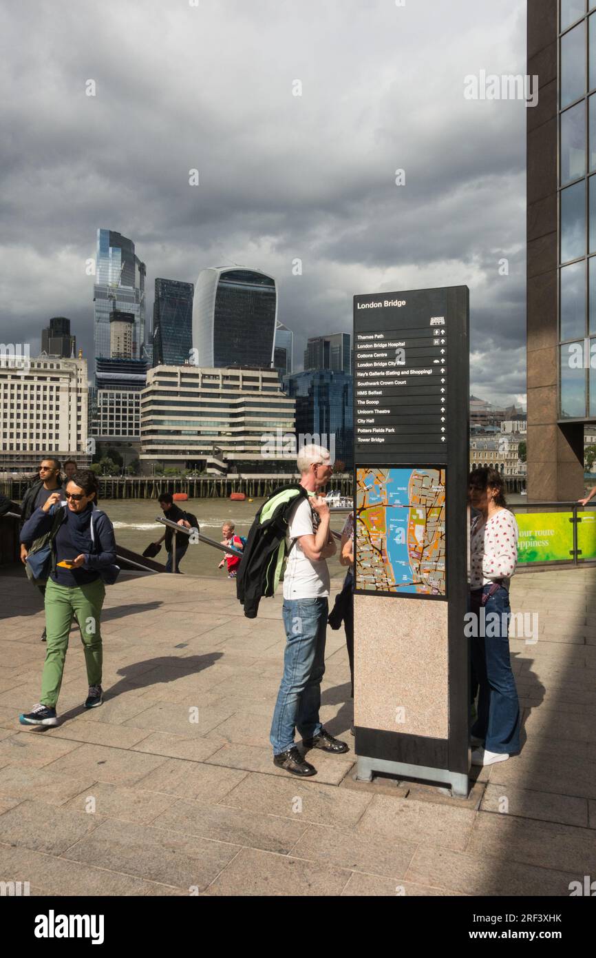 Storm clouds gathering over London Bridge directional signage and a middle-aged tourist viewing directional information, London, England, U.K. Stock Photo