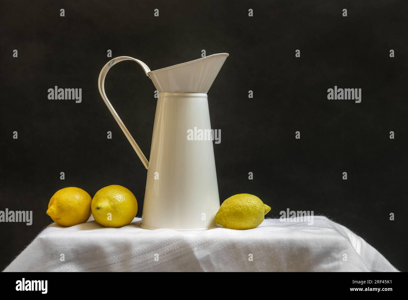 Cream enameled tin jug with a few loose ripe lemons on a white delicata cloth Stock Photo