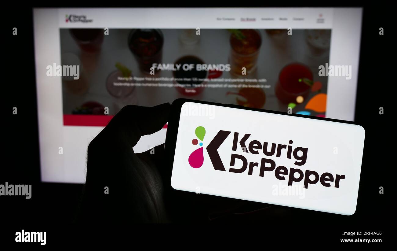 Person holding mobile phone with logo of American beverage company Keurig Dr Pepper Inc. on screen in front of web page. Focus on phone display. Stock Photo
