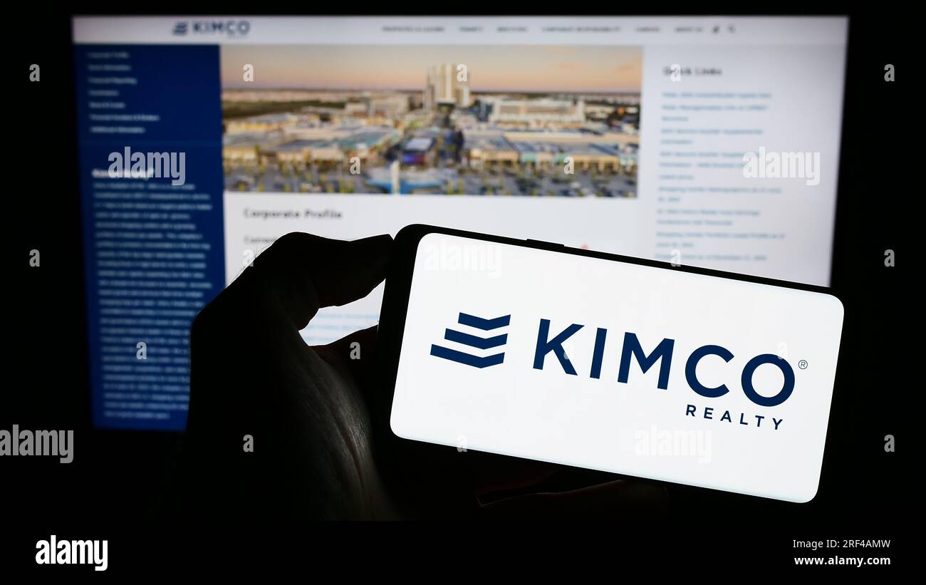 Person holding smartphone with logo of US real estate company Kimco Realty Corporation on screen in front of website. Focus on phone display. Stock Photo