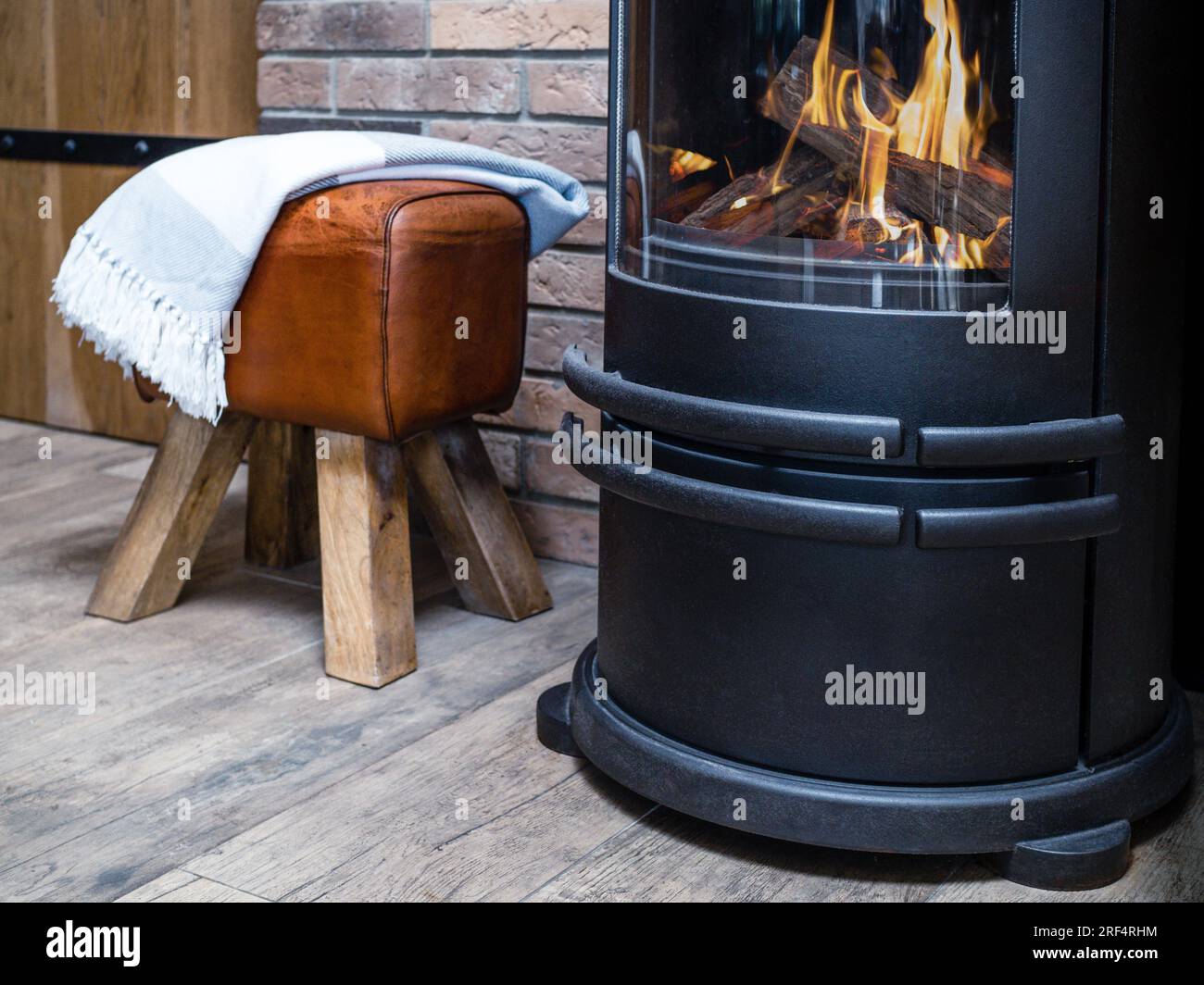 Cosy metal stove fireplace with burning flame behind a glass door and leather stool and blanket.  Stock Photo