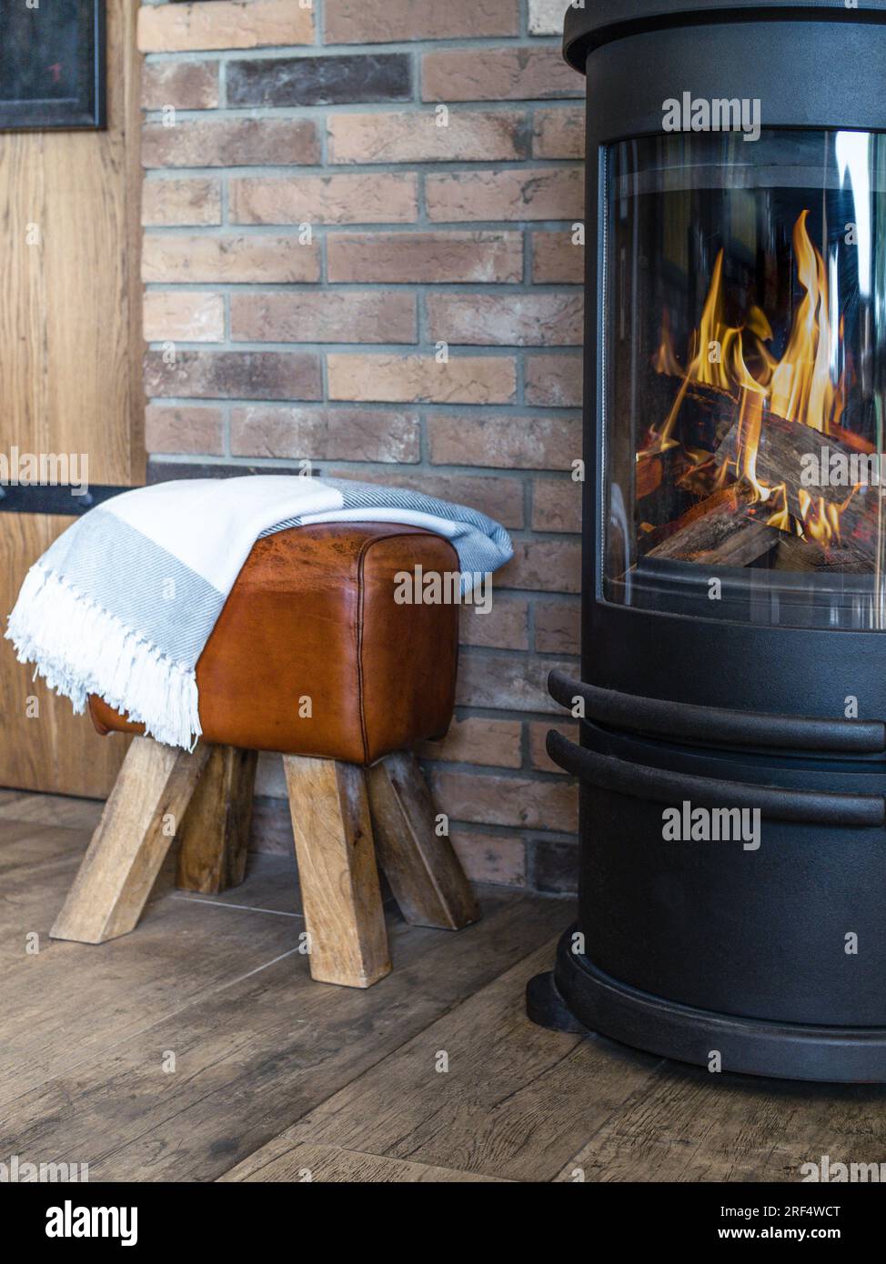 Cosy metal stove fireplace with burning flame behind a glass door and leather stool. Stock Photo