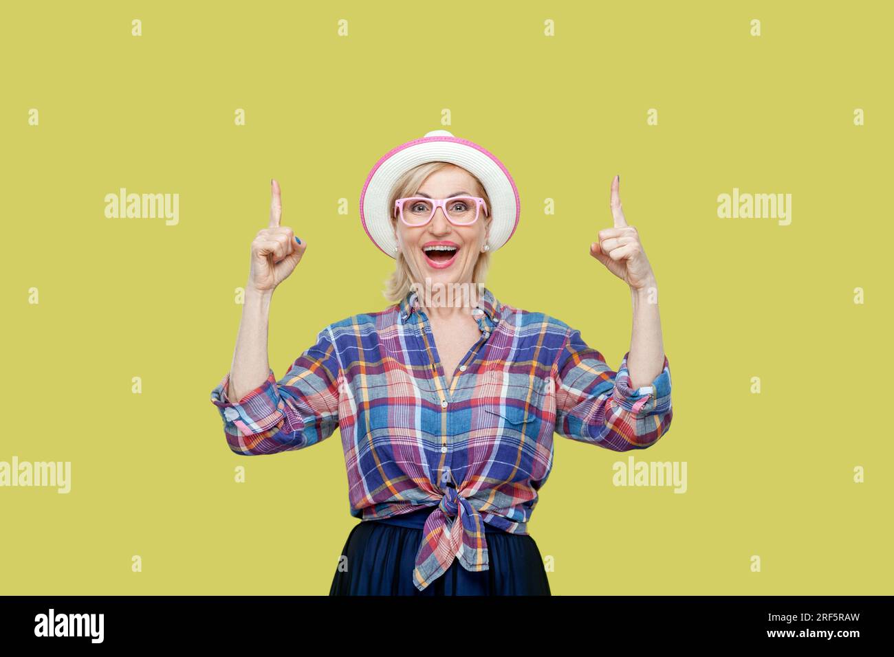 Senior woman wearing checkered shirt, hat and eyeglasses looks with widely opened mouth up, sees something amazed, indicates with fore fingers. Indoor studio shot isolated on yellow background. Stock Photo