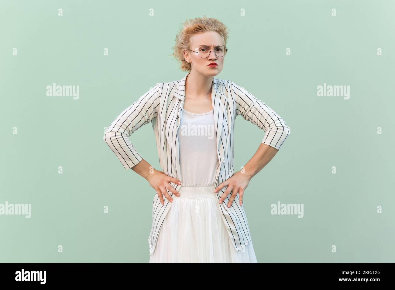 Portrait of strict bossy blonde woman wearing striped shirt and skirt looking at camera with serious expression, keeps hands on hips. Indoor studio shot isolated on light green background. Stock Photo