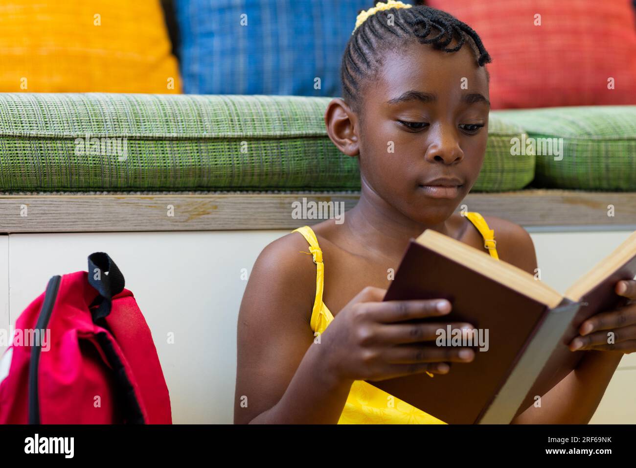 Focused african american schoolgirl reading book over couch with ...