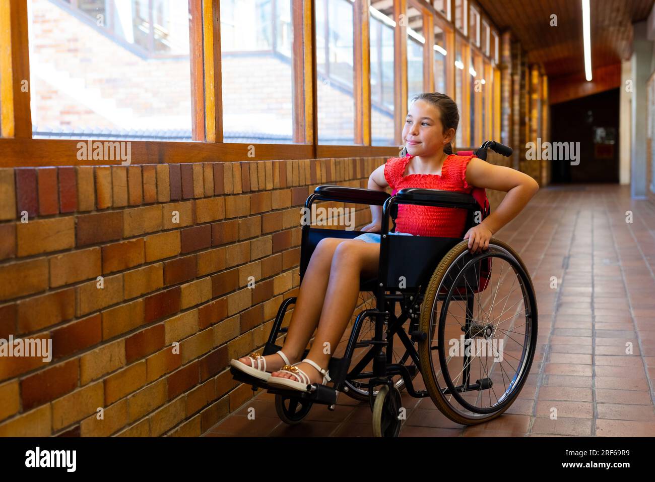 Happy caucasian schoolgirl sitting in wheelchair at elementary school corridor Stock Photo