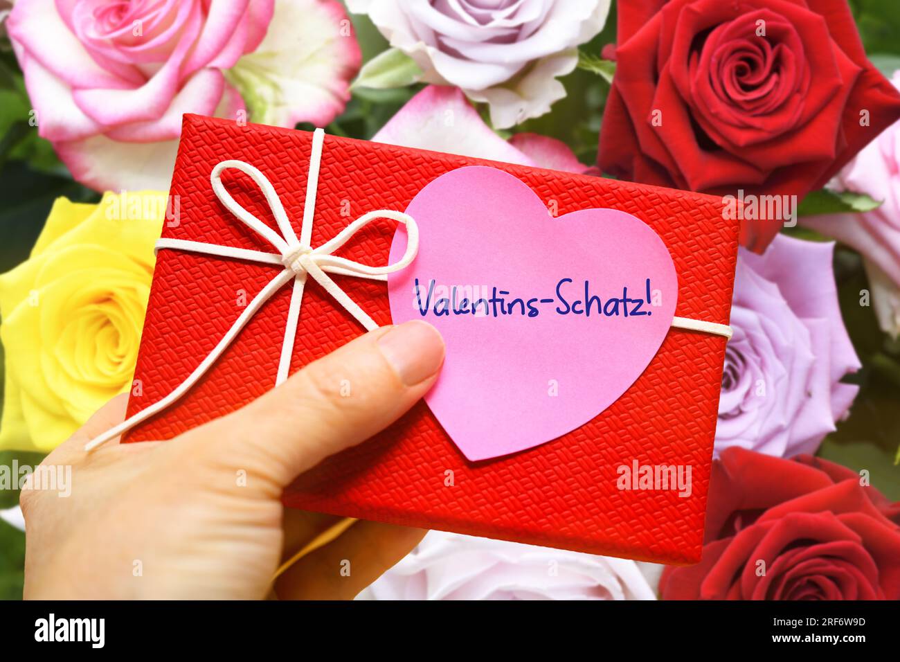 FOTOMONTAGE, Hand hält Geschenk mit einem herzförmigen Zettel und der Aufschrift Valentins-Schatz vor einem Blumenstrauß Stock Photo