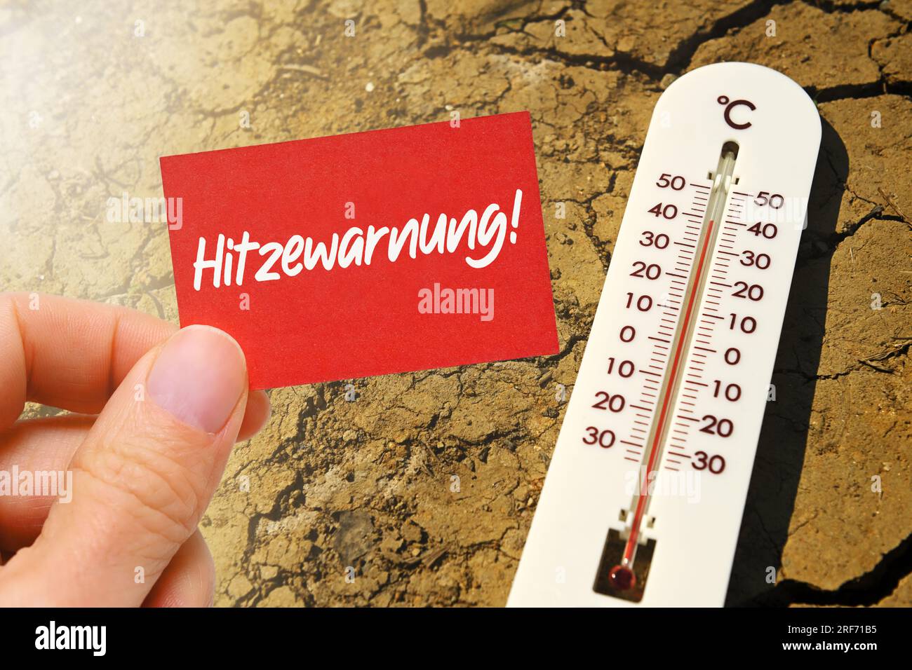 FOTOMONTAGE, Thermometer liegt auf gerissener Erde und Hand hält Schild mit Aufschrift Hitzewarnung Stock Photo