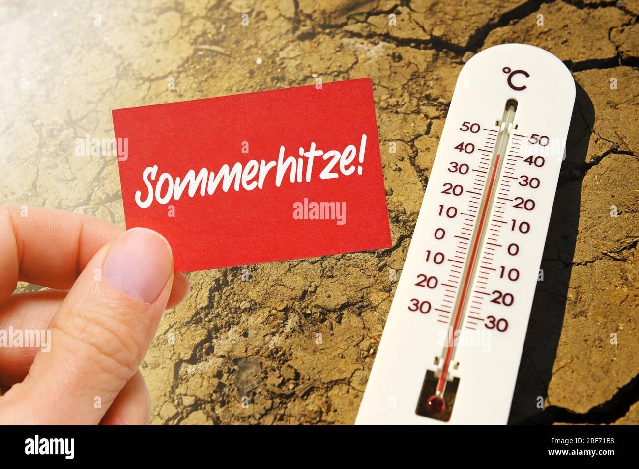 FOTOMONTAGE, Thermometer liegt auf gerissener Erde und Hand hält Schild mit Aufschrift Sommerhitze Stock Photo