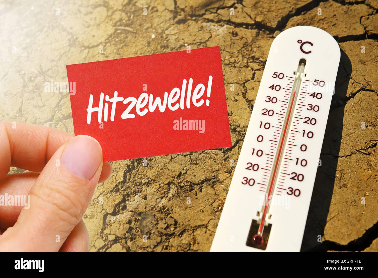 FOTOMONTAGE, Thermometer liegt auf gerissener Erde und Hand hält Schild mit Aufschrift Hitzewelle Stock Photo