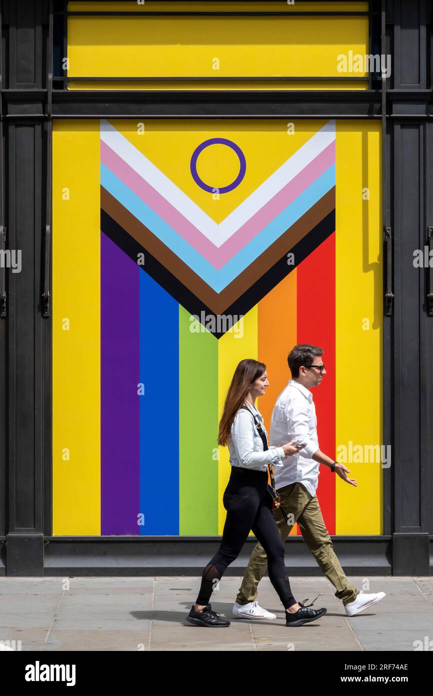 People pass a Pride Progress flag in an empty shop window on 9th July 2023 in London, United Kingdom. The flag includes the rainbow flag stripes to represent LGBTQ+ communities, with colors from the Transgender Pride Flag and to also represent people of colour. Stock Photo