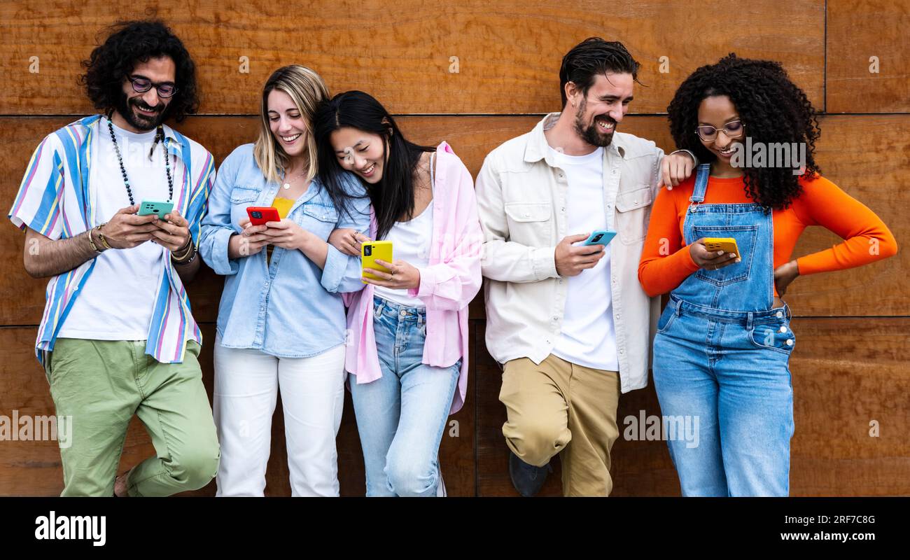 Diverse young confident people smiling and using their phones outside in a city while leaning against a wooden wall. Multiracial group of friends Stock Photo