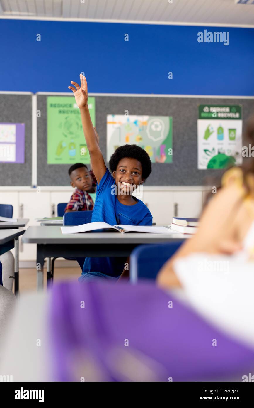 Diverse elementary schoolchildren sitting at desks and raising hands in ...