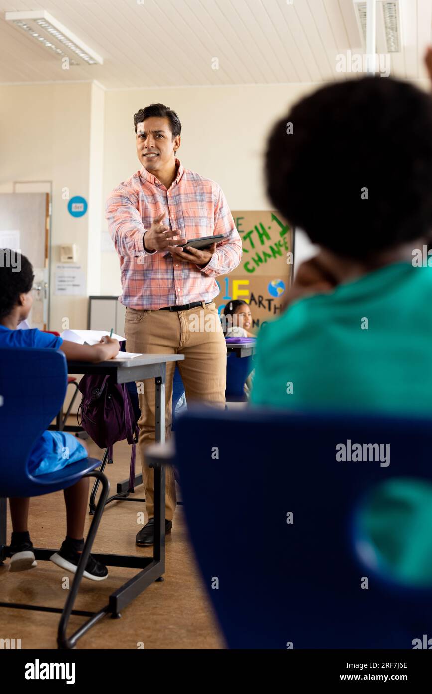 Diverse male teacher with tablet and elementary schoolchildren sitting ...