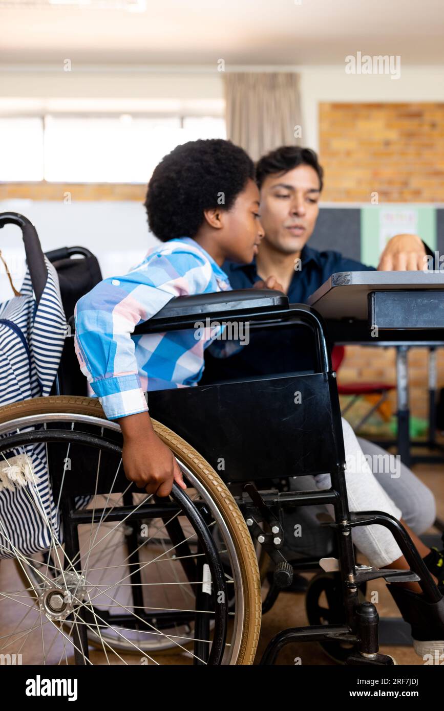 Diverse boy in wheelchair and male teacher in classroom at elementary school Stock Photo