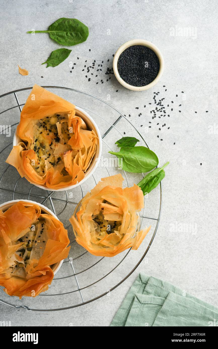 Filo pies with soft feta cheese and spinach in ceramic molds on old grey table background. Filo portions pies. Small Baked Spanakopita pies. Top view. Stock Photo