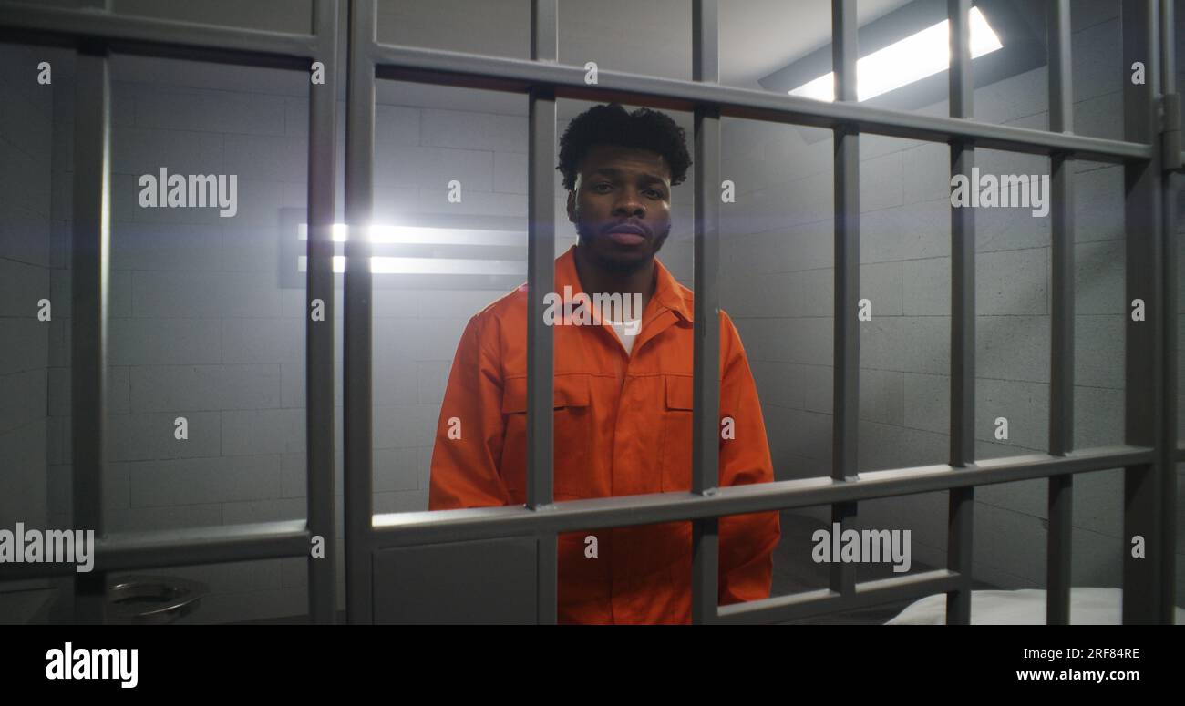 African American man in orange uniform leans hands on prison cell bars and looks at camera. Depressed prisoner serves imprisonment term in jail. Murderer in correctional facility or detention center. Stock Photo