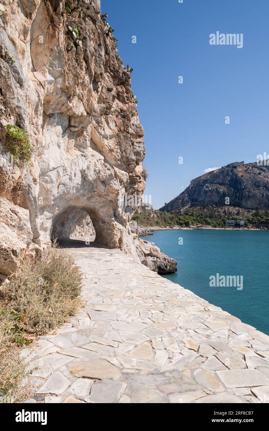 Photograph taken on the Arvanitia Walking Area, coastal path in Nafplio, with a wide paved path leading to a tunnel through the cliff, sea, beach and Stock Photo