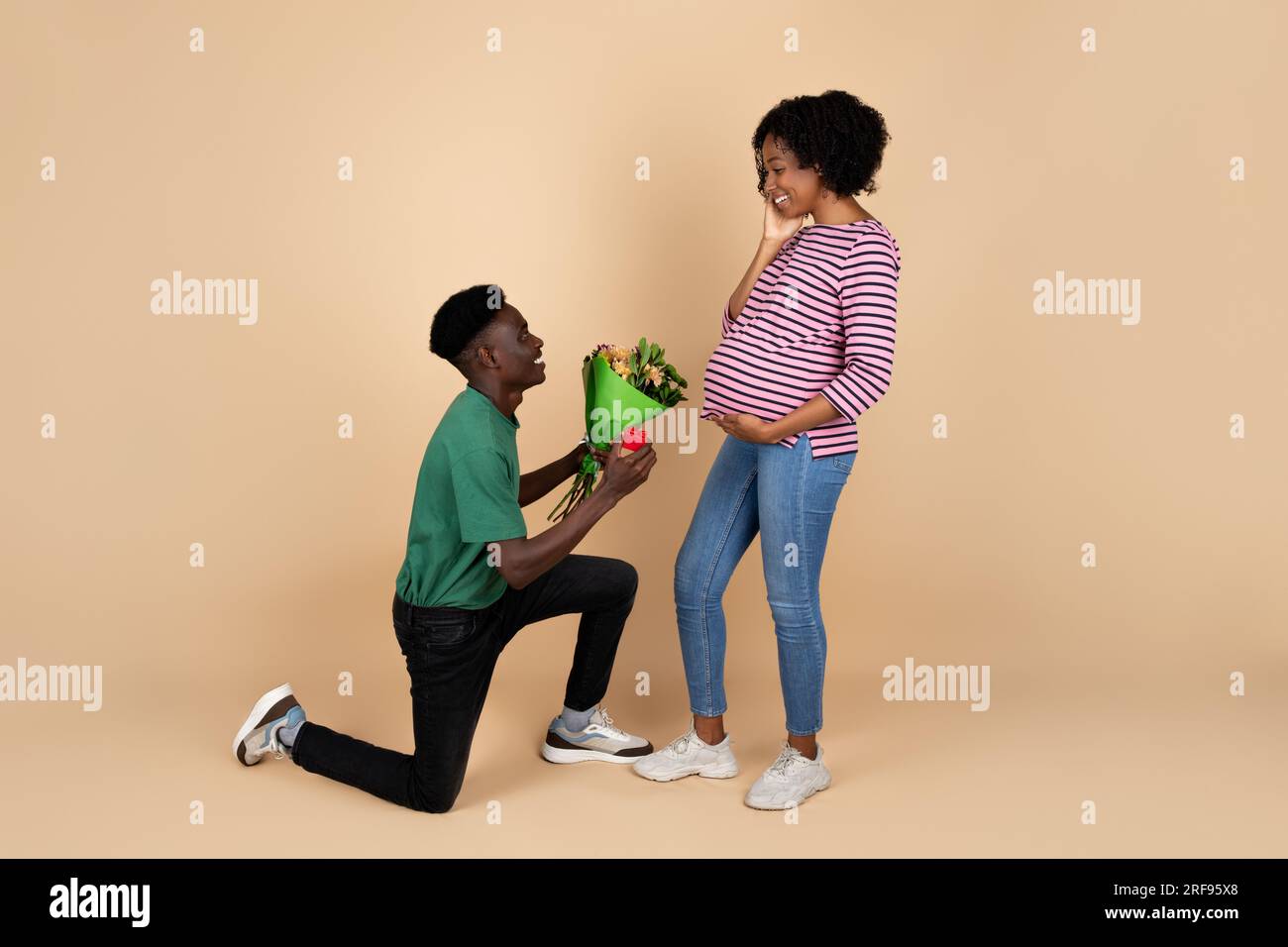Cheerful young black man kneeling, gives bouquet to lady with big belly, congratulations with pregnancy, expecting baby Stock Photo