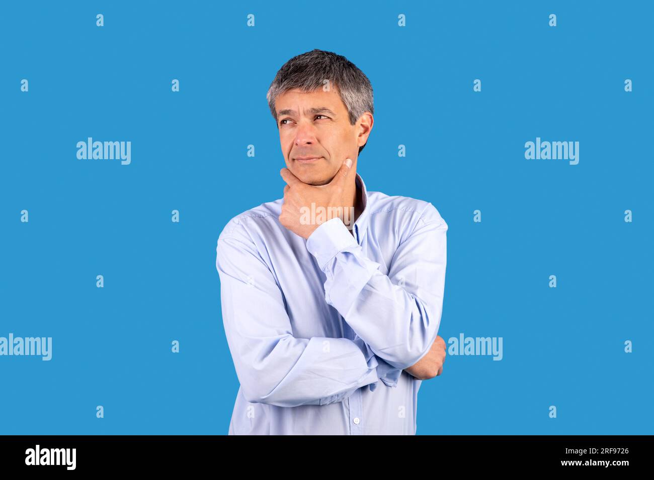 Pensive mature man touching chin with thoughtful expression, blue background Stock Photo