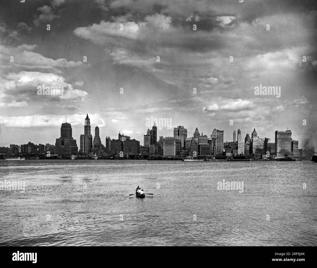 New York, New York:  c. 1927. Two men in a rowboat with an American flag for a bowsprit head off from New Jersey across the Hudson River towards Lower Manhattan. The Woolworth Building is the tallest at left, then next tallest to the right with the round dome is the Singer Building, and the double Equitable Building is to the south of that. Stock Photo